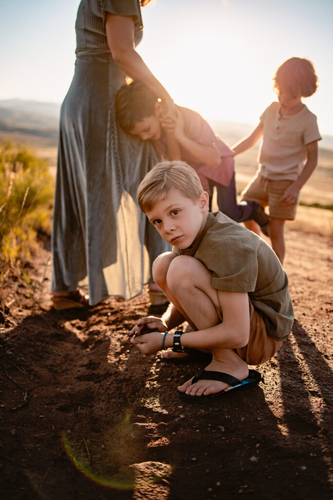 Little boy playing with the dirt while his brothers play with his mom in the background.