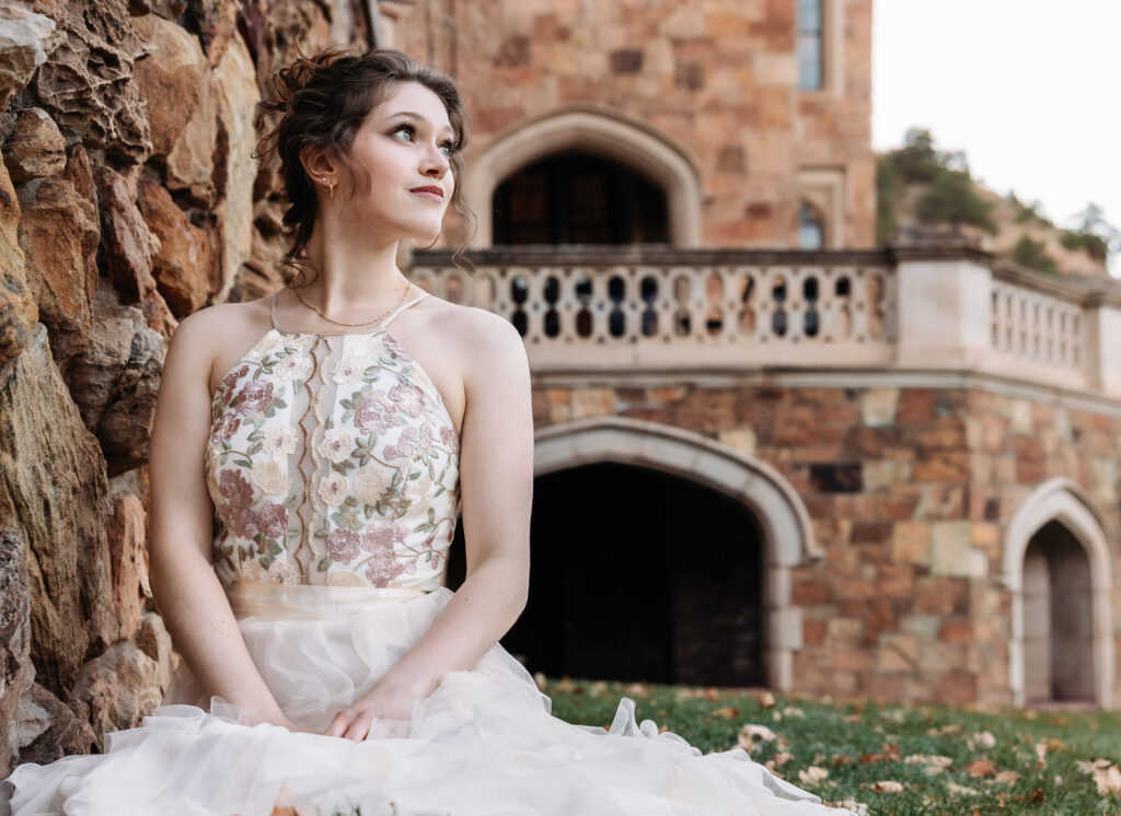 College senior sitting on the lawn in front of a castle wearing a beautiful gown.