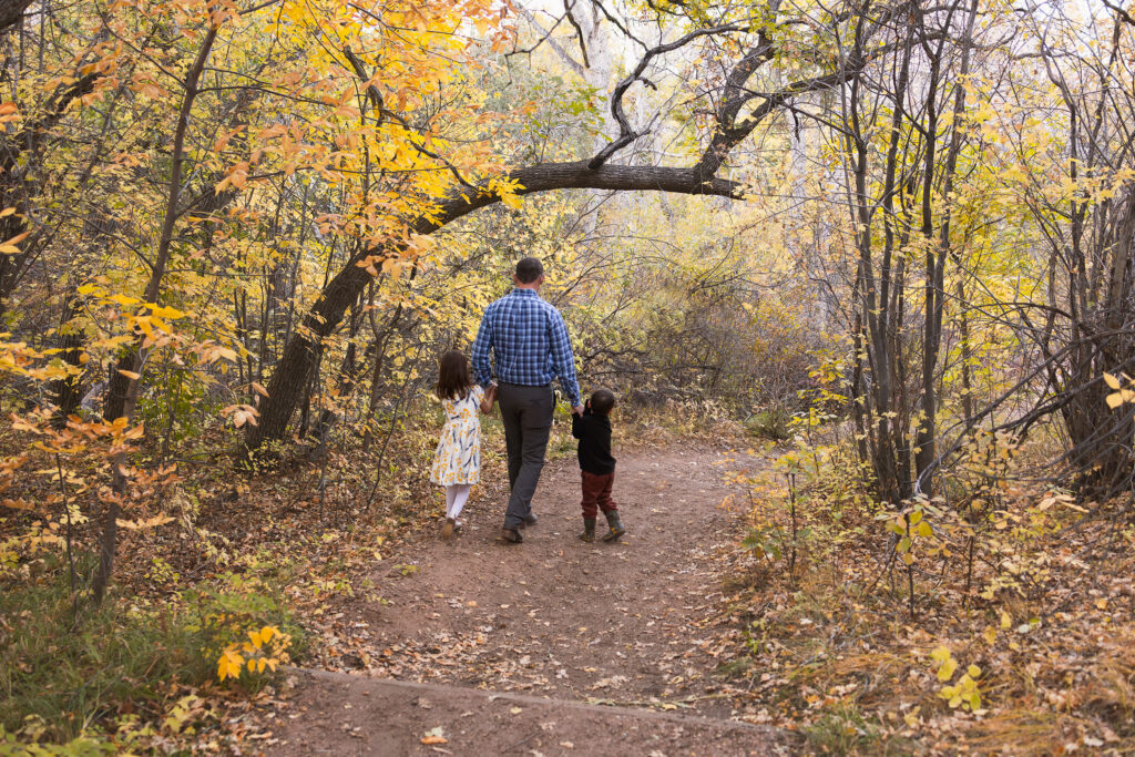 A father holding hands with his two children as he walks through a beautiful location with yellow leaves in the Fall in Colorado Springs.
