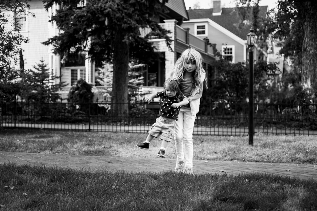 Big sister carries toddler little brother around the front yard at lifestyle family photography session in Colorado Springs, CO.