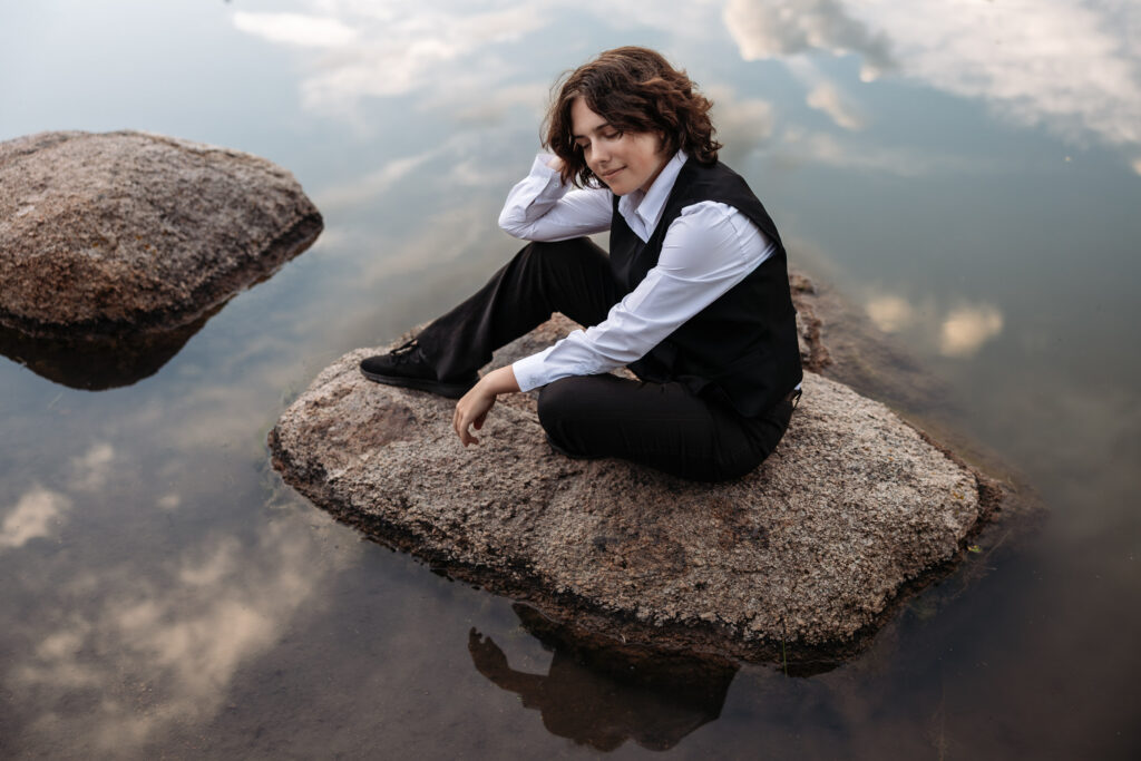 A high school senior posing on a rock with water from the lake reflecting a sky full of clouds location in Colorado Springs.