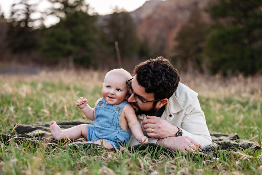 Colorado Springs baby photographer captures a Dad laying on a blanket while his six month old baby boy sits next to him. Baby has a big smile while Dad kisses baby's shoulder.