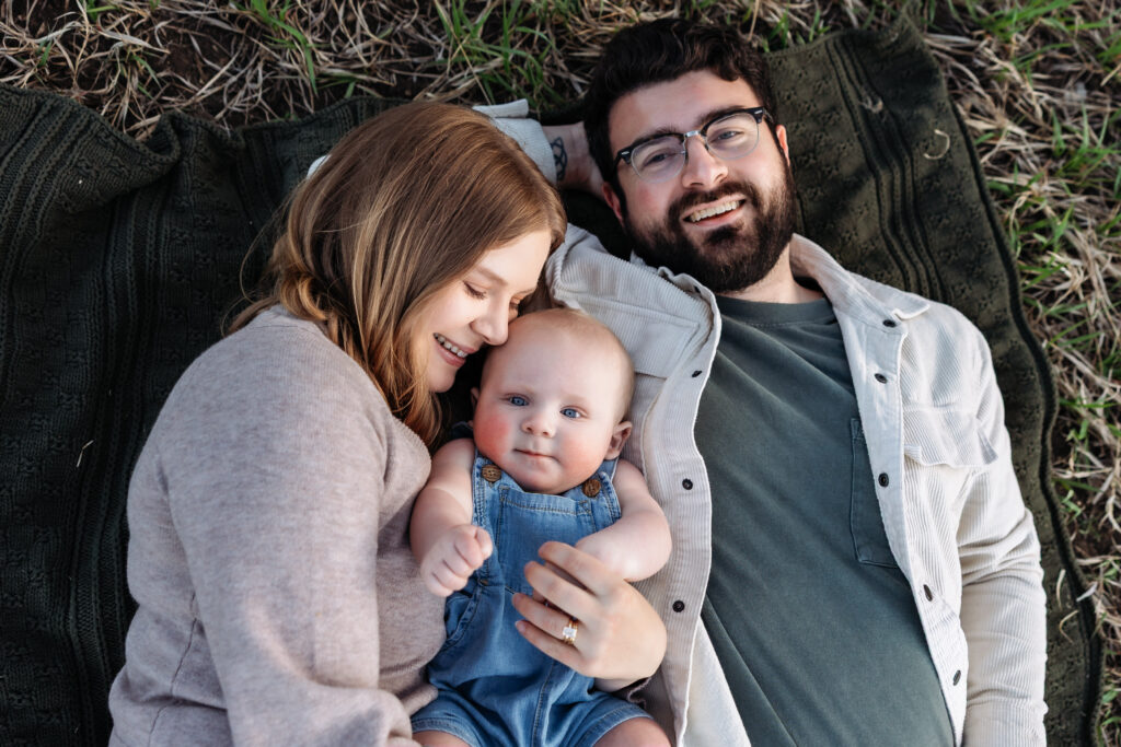 Young family with a six month old baby lay on a blanket at a baby photo session in Colorado Springs.
