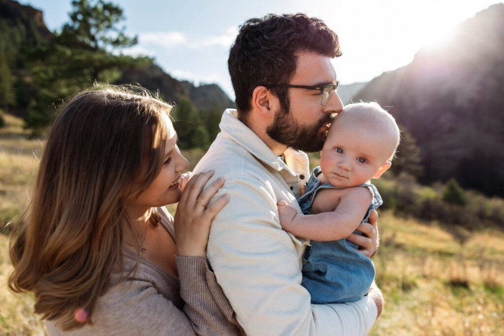 Young family of three at a baby photo session in Colorado Springs. Dad is holding 6 month old baby and kissing him on the forehead while mom smiles from behind.