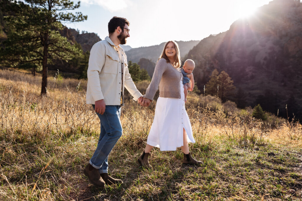 Mom and Dad hold hands while mom holds a baby and they walk in front of a beautiful mountain backdrop in Colorado Springs. Photo taken by at a baby photographer.