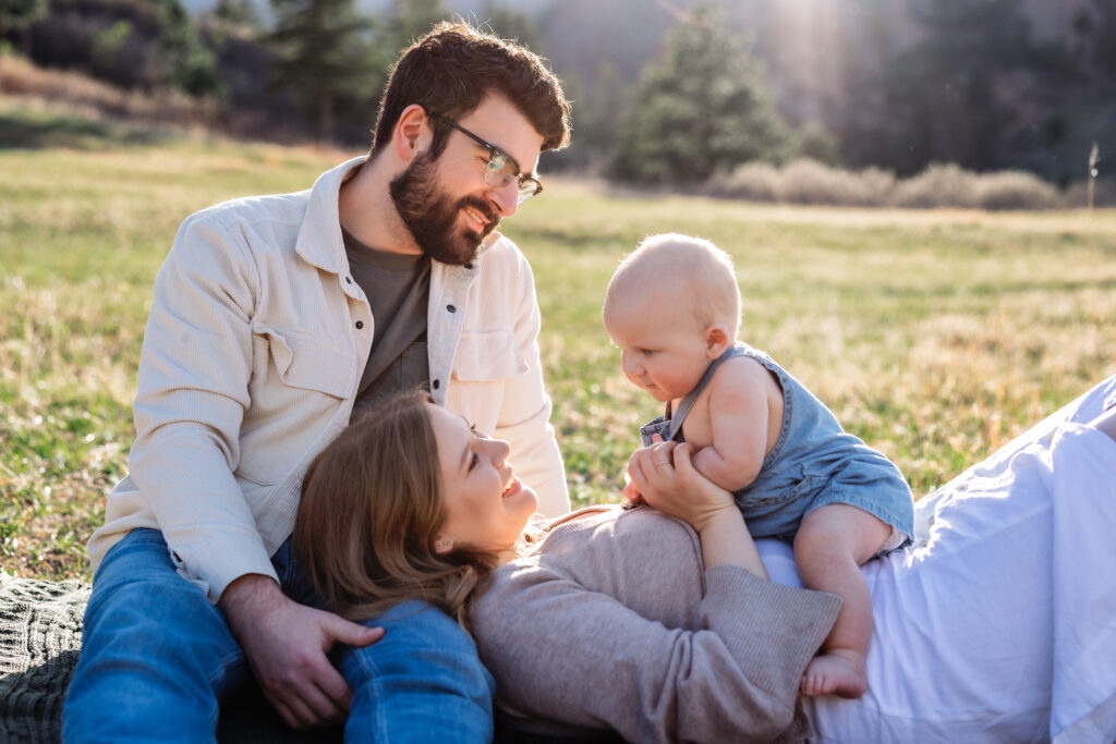 Six month old baby smiles at Mom while sitting on her stomach. Mom is laying on Dad's lap at a baby photo session in Colorado Springs.